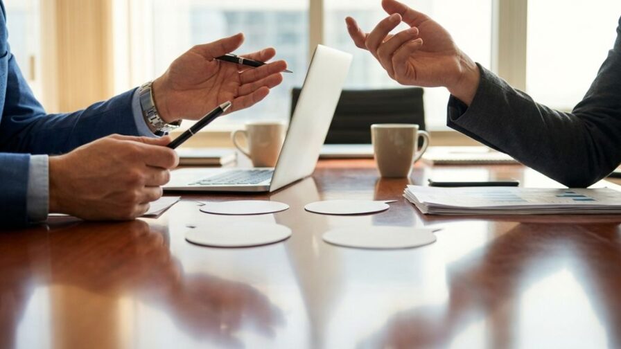 Twee professionele handen in gesprek boven vergadertafel, één met pen, kantooromgeving met laptop op achtergrond