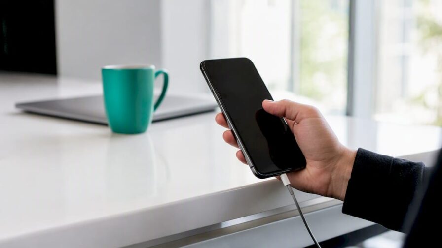 Een hand houdt een smartphone met een zwart scherm boven een donker bureaublad vast.